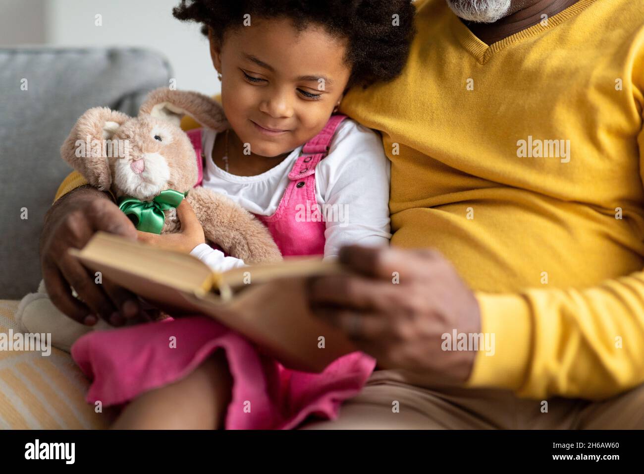 Lächelndes kleines afroamerikanisches Mädchen mit Spielzeug liest ein Buch mit einem älteren Großvater auf dem Sofa in der Inneneinrichtung, abgeschnitten, aus nächster Nähe. Studieren, Märchen togeth Stockfoto