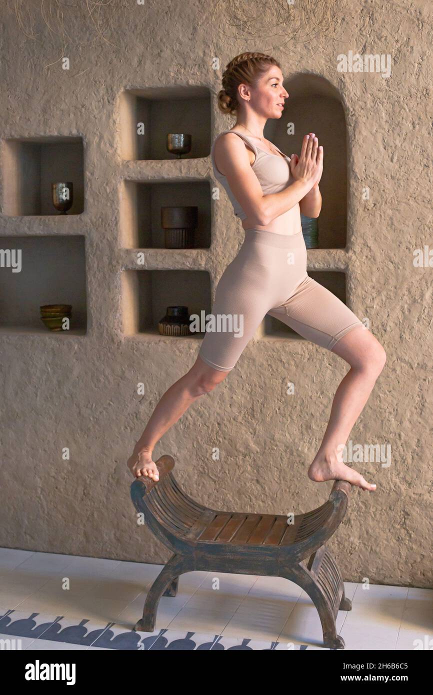 Eine schlanke, schöne Frau in einem beigen Oberteil und Leggings steht in einer Yoga-Pose auf einem Stuhl Stockfoto