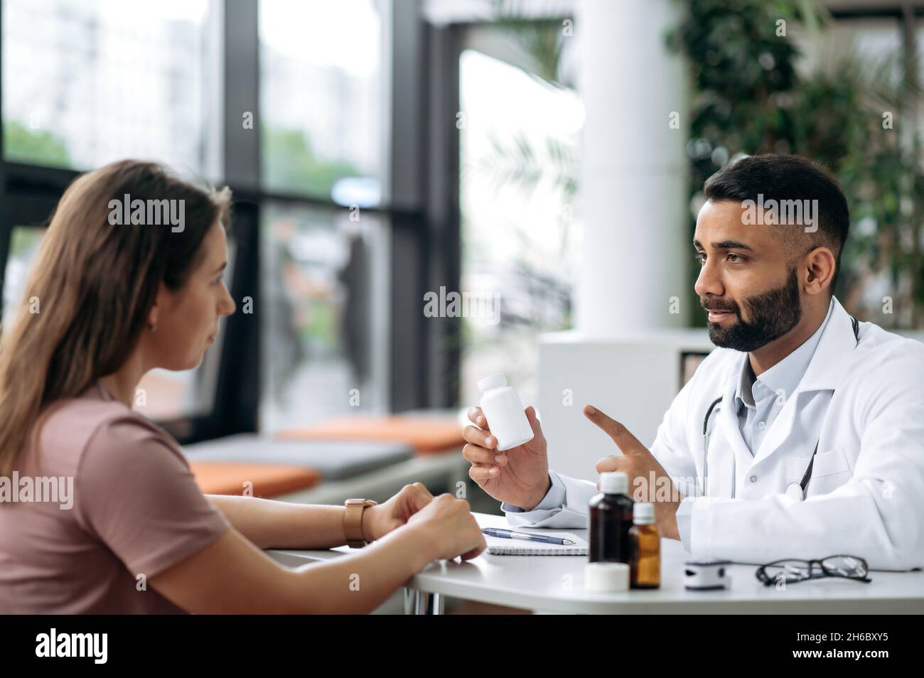 Konsultation mit einem Arzt in einer Klinik. Indischer Arzt der Allgemeinmedizin in einer medizinischen Uniform, berät den Patienten, verschreibt eine Behandlung, hört auf Beschwerden, gibt Ratschläge, Zeigt Medikamente an Stockfoto