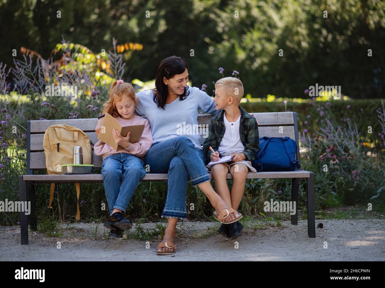 Glückliche ältere Frau mit Enkelkindern, die auf der Bank sitzen und bei Hausaufgaben im Park helfen. Stockfoto
