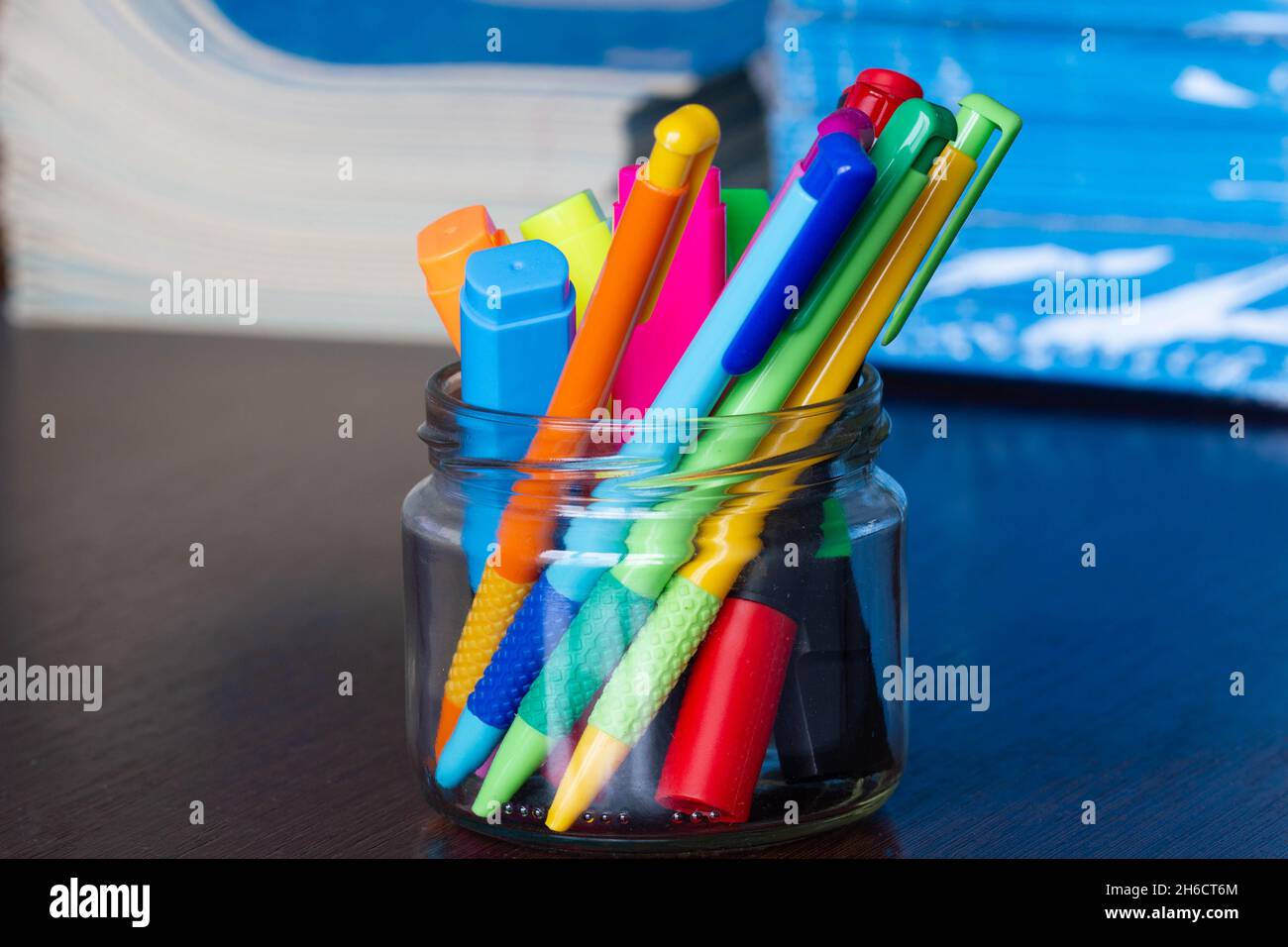 Mehrfarbige Stifte und Marker in einem Standfuß auf dem Hintergrund des Stapels von Notizbüchern. Eine Packung blauer Schulnotizbücher auf dem Tisch im Klassenzimmer. Stapel Stockfoto