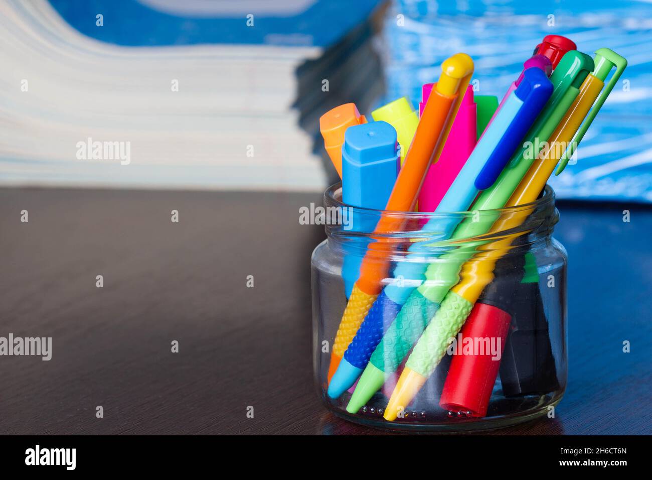 Mehrfarbige Stifte und Marker in einem Standfuß auf dem Hintergrund des Stapels von Notizbüchern. Eine Packung blauer Schulnotizbücher auf dem Tisch im Klassenzimmer. Stapel Stockfoto