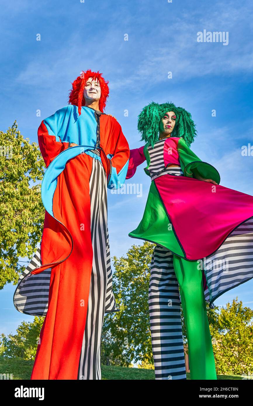 Von unten lustige Showdarsteller auf Stelzen in bunten Clownkostümen und Perücken, die im grünen Park und während der festlichen Veranstaltung zusammen stehen Stockfoto