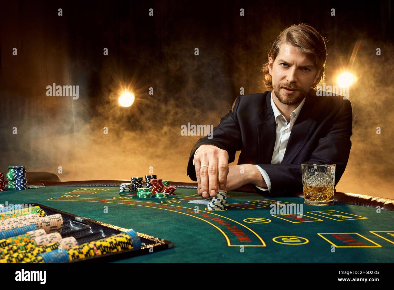 Ein Mann in einem Geschäftsanzug, der am Spieltisch sitzt. Männlicher Spieler. Leidenschaft, Karten, Chips, Alkohol, Würfel, Glücksspiel, Casino - es ist wie männliche Unterhaltung. Stockfoto