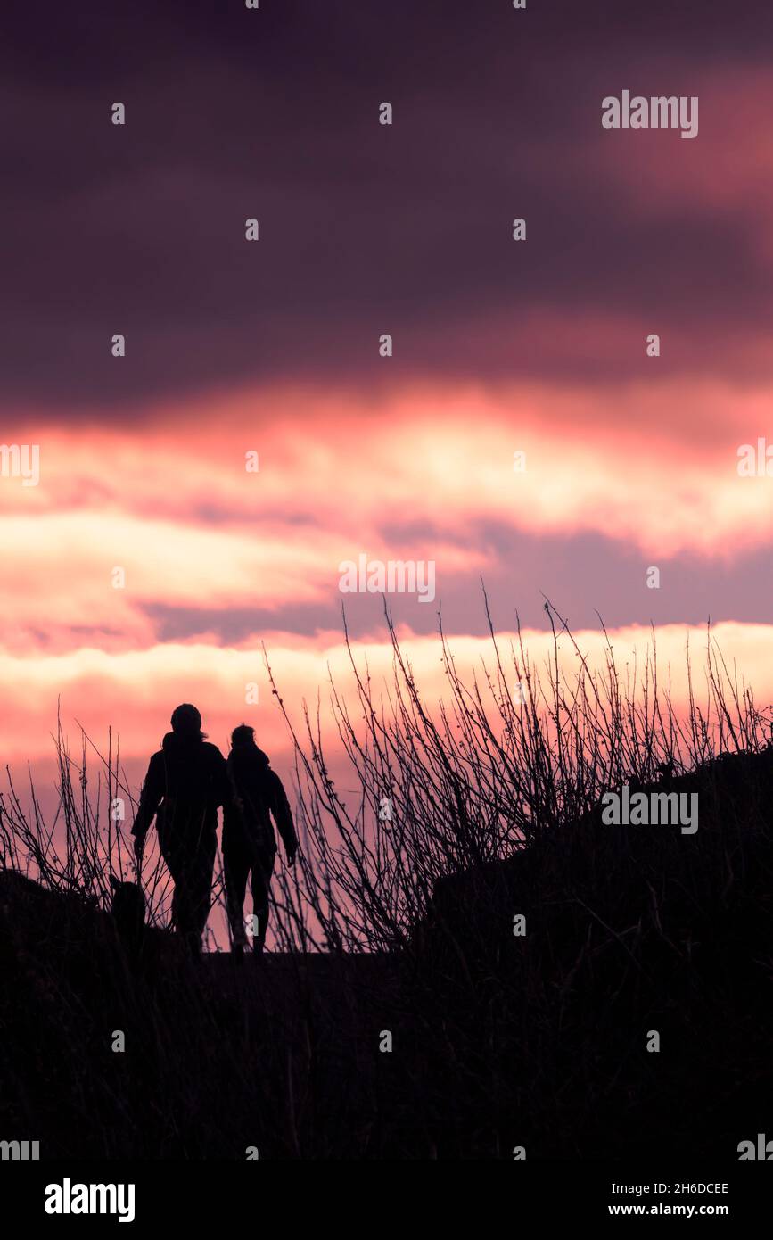 Ein Paar, das am Ende des Tages am Fistral in Newquay in Cornwall den Küstenpfad entlang geht, der von einem bunten Himmel umraunt wird. Stockfoto
