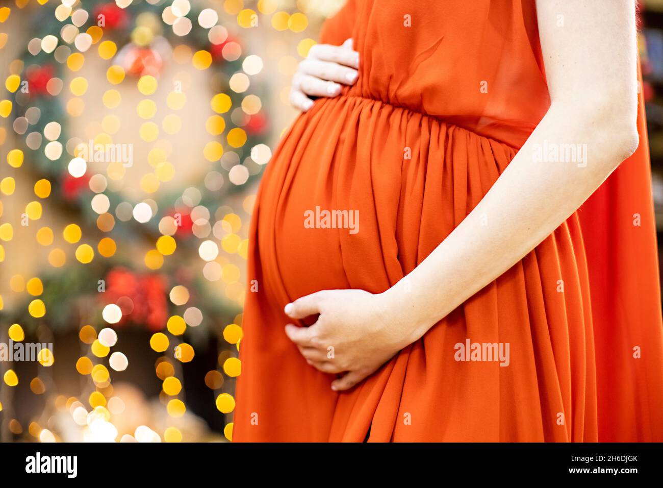 Bestes weihnachtsgeschenk. Schwangere in einem roten Kleid umarmt ihren Bauch vor dem Hintergrund des Weihnachtswohnzimmers in Unschärfe. Nahaufnahme kein Gesicht. Stockfoto