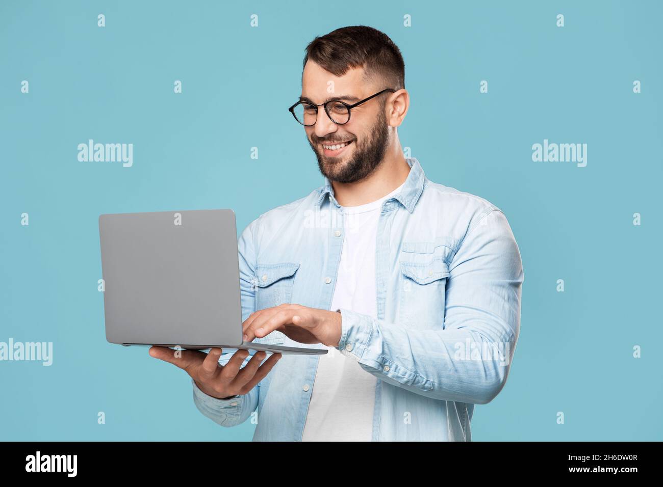 Lächelnd attraktiver erwachsener europäischer Mann in Brille, der am Computer tippt, funktioniert ferngesteuert, isoliert auf blauem Hintergrund Stockfoto