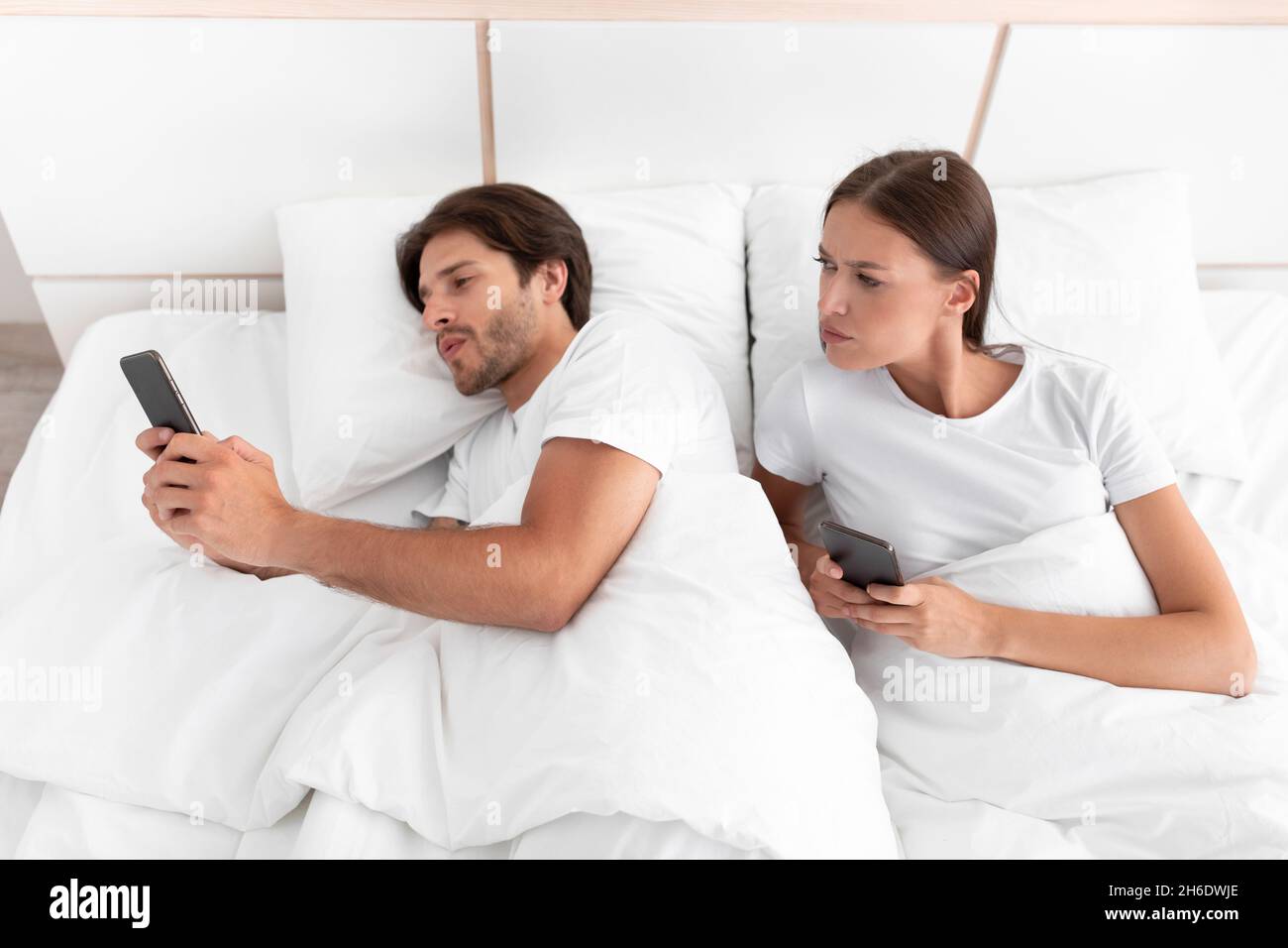 Verärgerte misstrauische junge europäische Frau schaut auf das Telefon des Ehemanns, Kerl, der in sozialen Netzwerken im Bett chattet Stockfoto