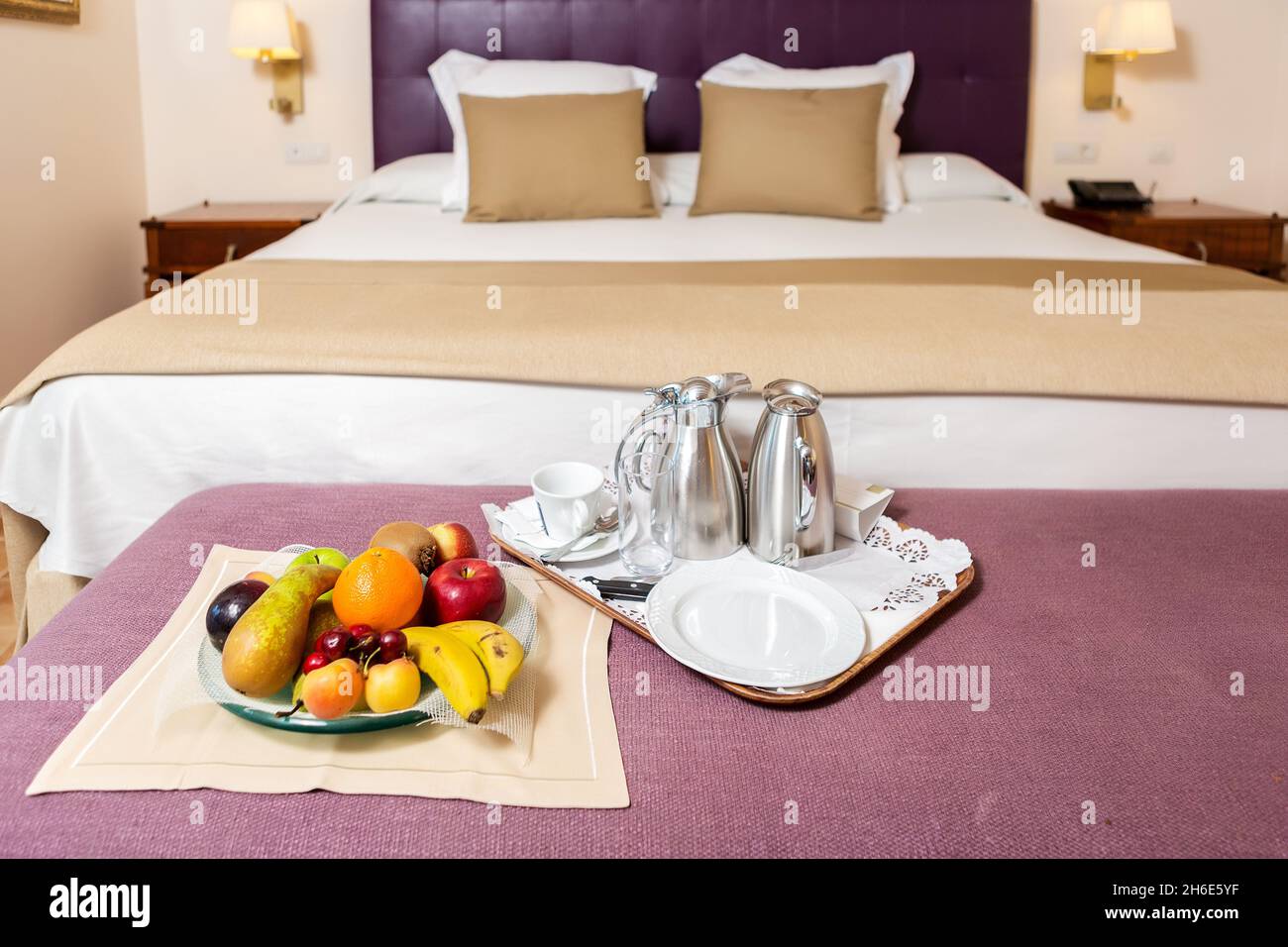 Teller mit verschiedenen Früchten und Kaffee-Set auf Tablett platziert Auf dem Tisch mit violetter Abdeckung auf dem Hintergrund des Bettes in Weiße und braune Farben Stockfoto