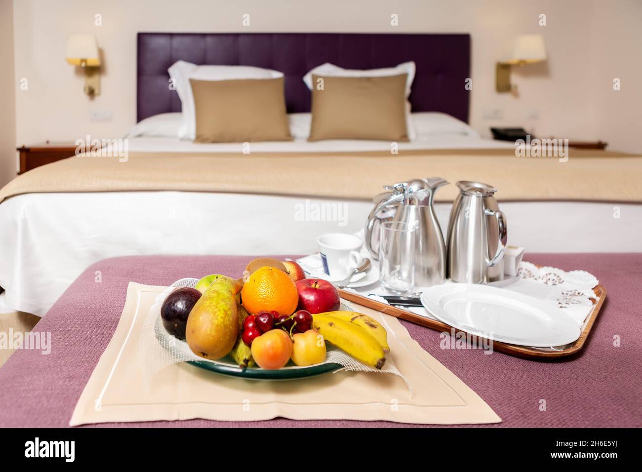 Teller mit bunten frischen Früchten und Kaffee-Set auf Tablett Stellen Sie auf den Tisch mit violetter Abdeckung auf dem Hintergrund des Bettes Mit weißem und braunem Bettlaken Stockfoto