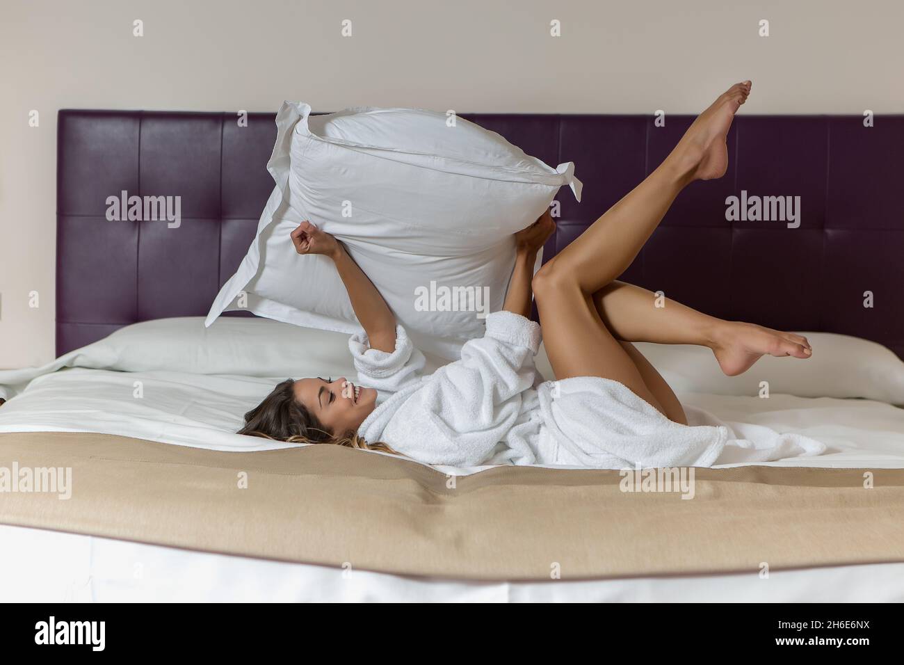 Seitenansicht einer lächelnden jungen Frau mit Kissen in den Händen, die morgens im Hotelbett schwelgen Stockfoto