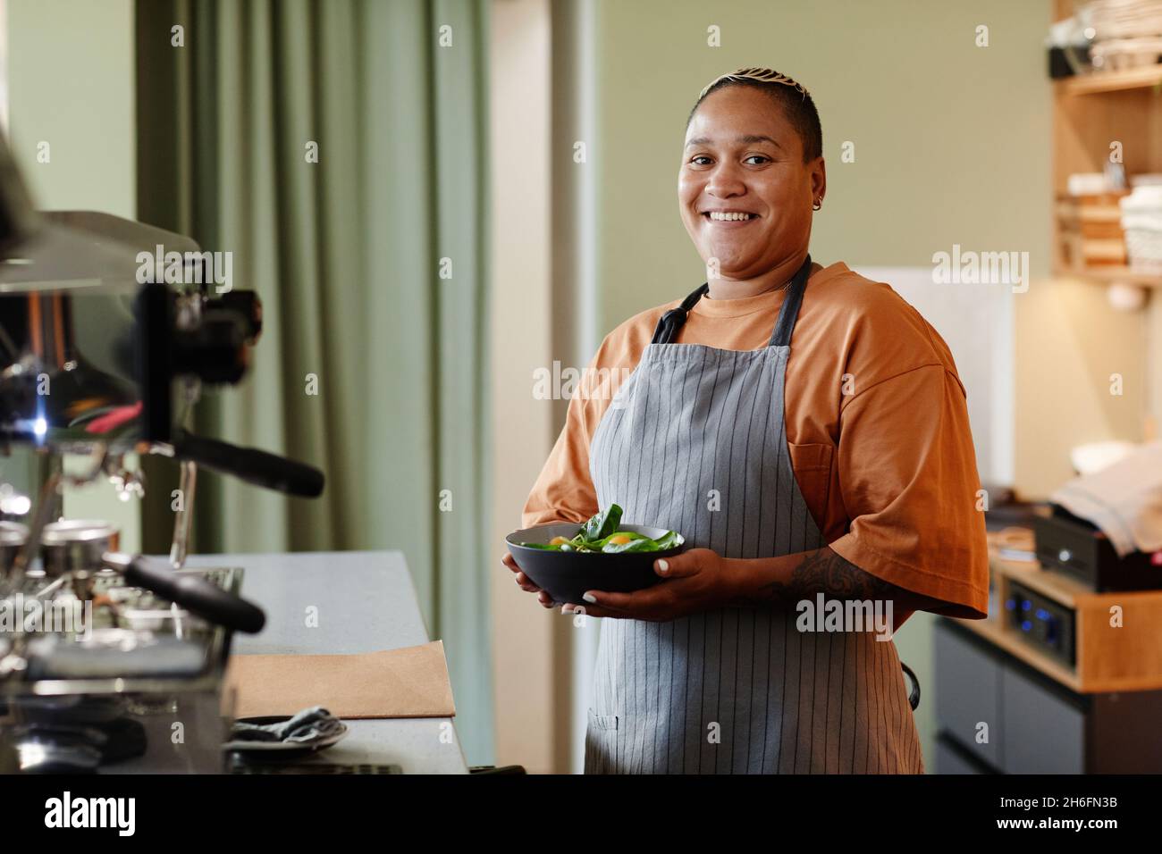 Horizontales mittleres Porträt der fröhlichen jungen afroamerikanischen Frau mit stilvollen kurzen Haaren trägt Schürze stehend in Café Küche mit Schüssel mit s Stockfoto