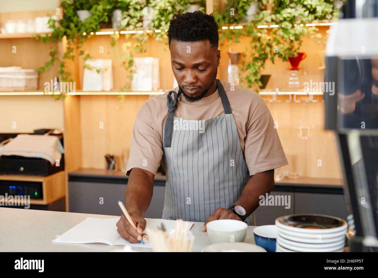 Horizontales mittleres Porträt eines jungen schwarzen Mannes mit Schürze, der an der Küchentheke des Cafés steht und etwas in einem Notizbuch schreibt Stockfoto