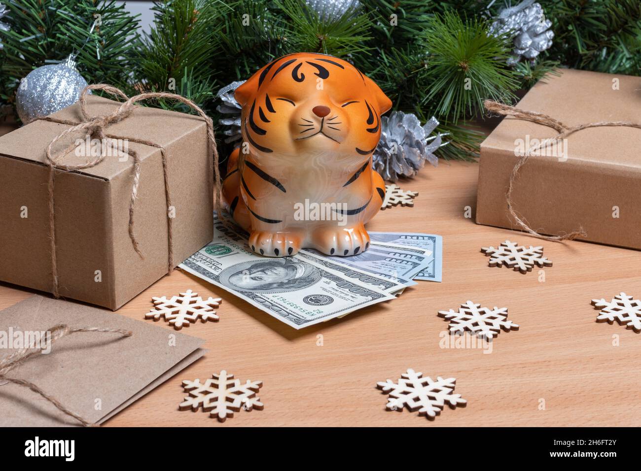 Ein Sparschwein in der Form eines Tigers, der auf Gelddollar auf einem hölzernen Hintergrund mit Geschenkschachteln, Schneeflocken und Tannenzweigen steht. Erfolg und Wohlstand Stockfoto