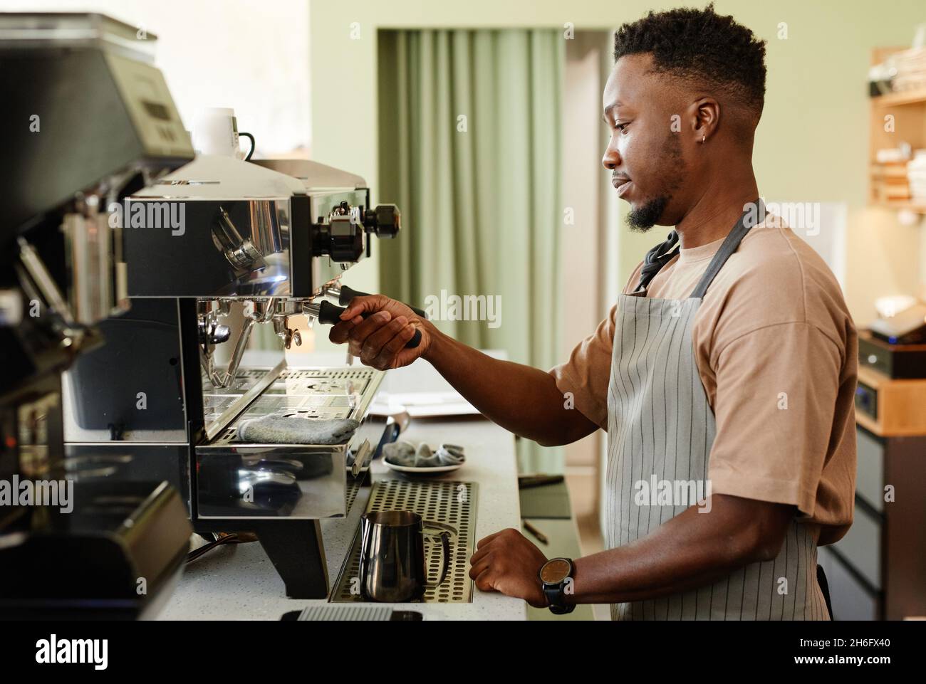 Horizontale Seitenansicht des hübschen jungen afroamerikanischen Mannes, der als Barista in einem kleinen Café arbeitet und Espresso mit einer modernen Kaffeemaschine zubereitet Stockfoto