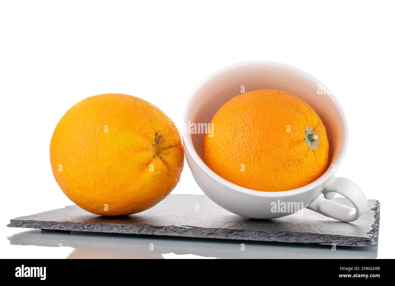 Zwei organische, leuchtend gelbe Orangen mit einer weißen Tasse auf einem Schiefertablett, Nahaufnahme, isoliert auf Weiß. Stockfoto