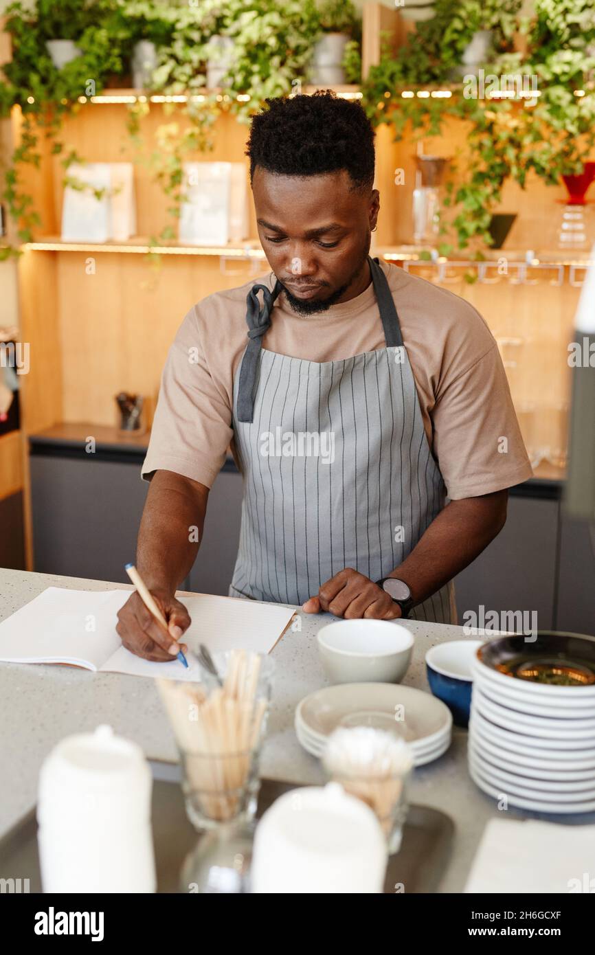 Vertikales mittleres Porträt eines jungen afroamerikanischen Mannes mit Schürze, der am Küchentisch des Cafés steht und etwas in einem Notizbuch schreibt Stockfoto