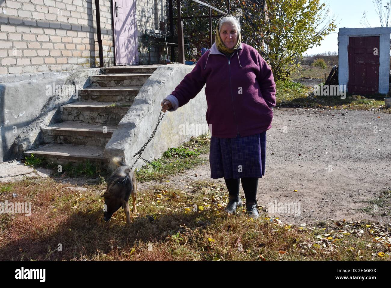 Eine ältere Dame steht mit ihrem Hund im Hinterhof ihres Hauses am Rande des Dorfes Werknotoretske. Werchnotoretske ist eine ukrainische Siedlung, die sich direkt an der Front im Bezirk Jassynuvata im Bezirk Donezk im Osten der Ukraine, 24, 2 km vom Zentrum der Stadt Donezk (DPR) entfernt befindet. Während des Krieges im Donbass, der Mitte April 2014 begann, wurde in der Nähe der Siedlung die Trennungslinie zwischen kriegführenden Parteien errichtet. Das Dorf war bis Ende 2017 eine Grauzone. Zur Zeit ist Werchnotoretske ein Territorium der Ukraine. Der Konflikt hat es gebracht Stockfoto