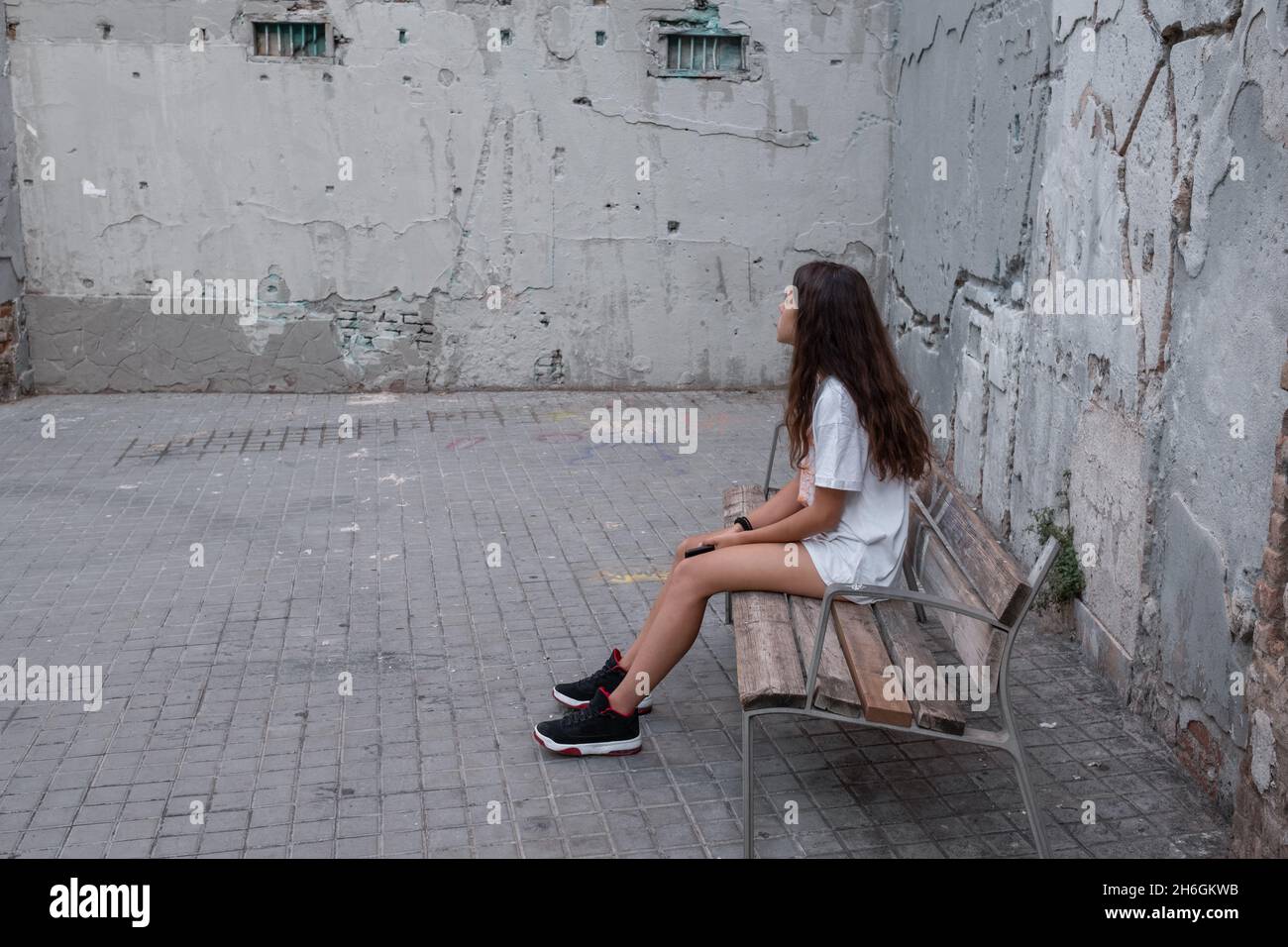 Porträt eines Teenagers, das auf einer Bank sitzt und ein Smartphone in den Händen hält. Sie schaut in die Richtung der schmutzigen Wand. Stockfoto