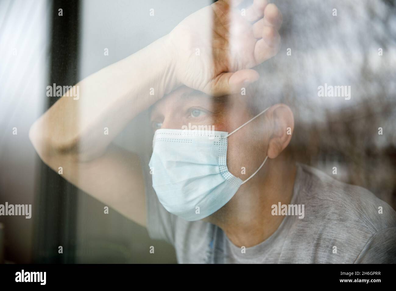 Mann mit Gesichtsmaske, der in einer Quarantäne sitzt und durch das Fenster schaut. Stockfoto