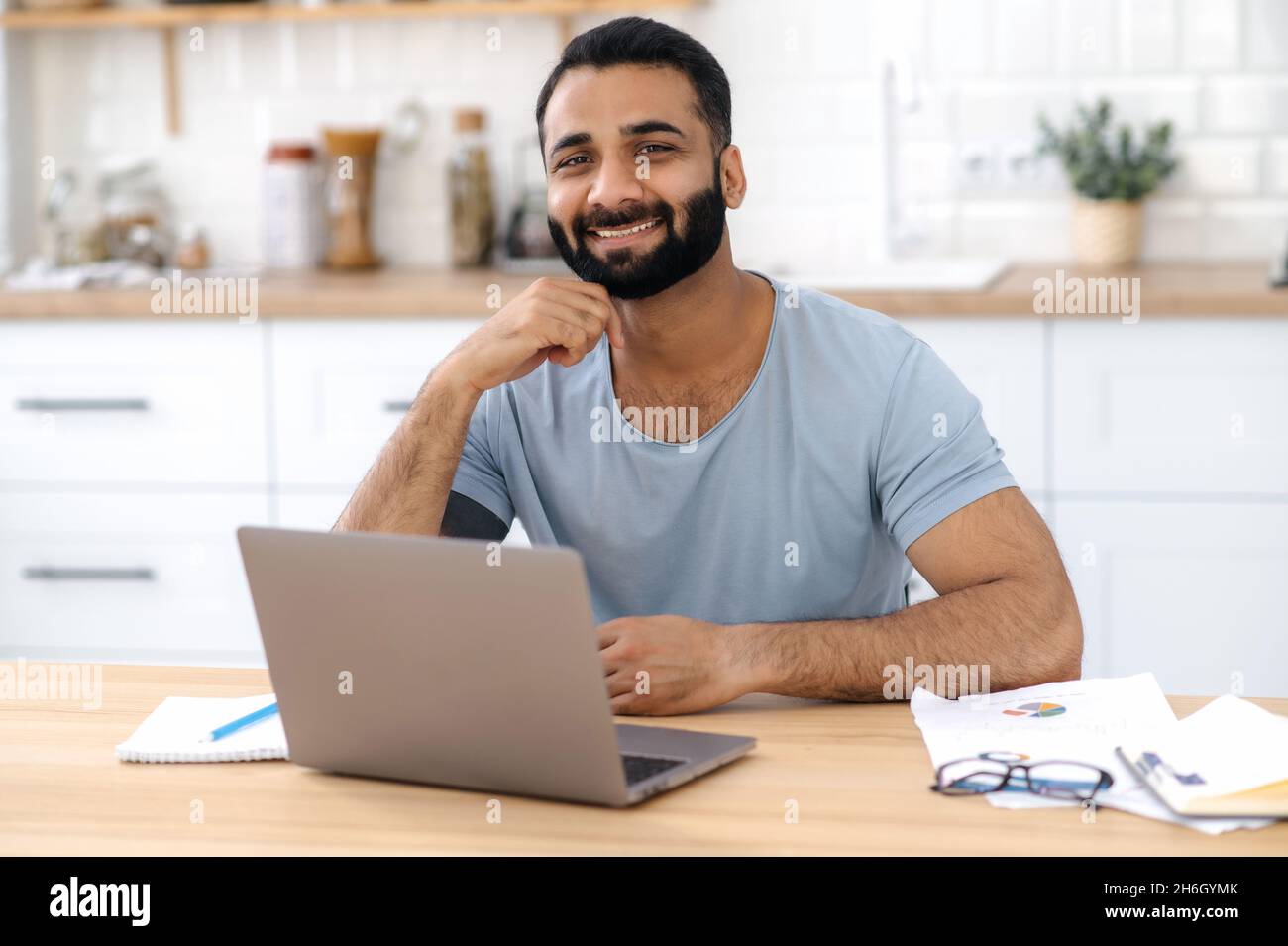 Porträt eines charismatischen, positiven jungen erwachsenen Inders, Freiberuflers, Designers oder IT-Spezialisten, arbeitet von zu Hause aus mit einem Laptop, blickt direkt in die Kamera und lächelt freundlich Stockfoto