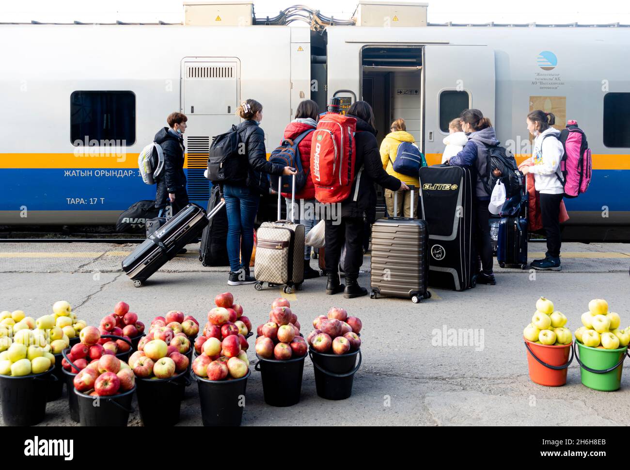 Passagiere, die an einer Bahnhaltestelle in einen Zug einsteigen. Eimer mit frischen Herbstäpfeln werden auf den Boden gestellt und von lokalen Händlern verkauft. Almaty, Kasachstan Stockfoto