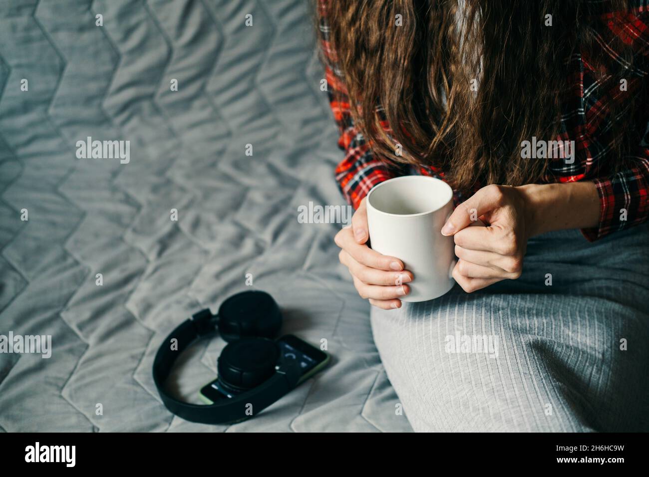 Ein Mädchen sitzt auf einem grauen Sofa, mit einer Tasse Kaffee in den Händen, einem Telefon und kabellosen Kopfhörern, Copy Space Freizeit zu Hause. Liebe zur Musik. Stockfoto