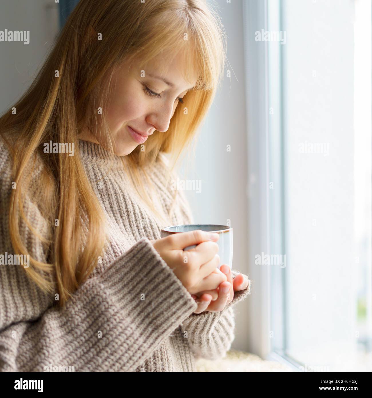 Fröhliche Frau mit einer Tasse heißem Getränk, die neben dem Fenster im Pullover steht. Ein langsamer Lebensstil Stockfoto
