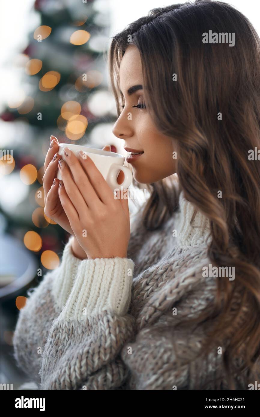 Seitenansicht der schönen Frau in Strickpullover genießen heiße Getränke in gemütlichen Hause. Verwischen Sie den Hintergrund des weihnachtsbaums. Entspannung während der Winterferien. Stockfoto