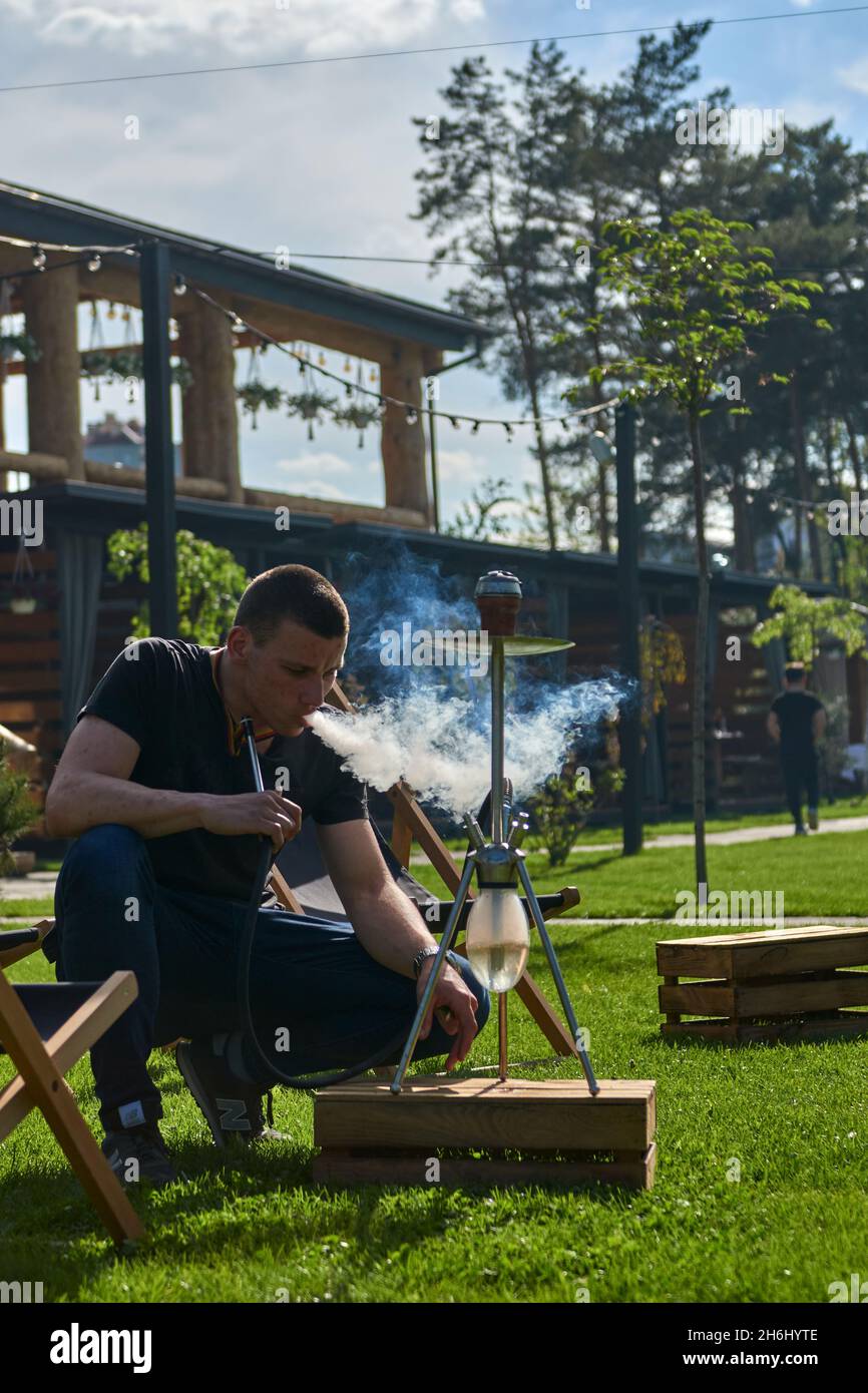 Mann, der an einem warmen sonnigen Tag Shesha, Shisha-Shisha in der Natur raucht. Östlicher Shisha Rauchen, aktive Erholung an der frischen Luft. Liegestühle auf geen Gras Stockfoto