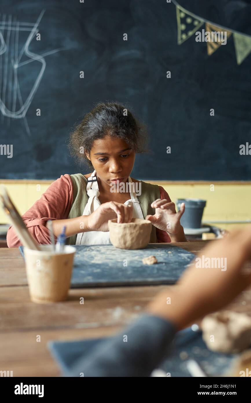 Nettes jugendliches Mädchen in Schürze, das Tonschüssel oder Becher macht, während es am Tisch gegen die Tafel bei der Lektion der Handarbeit sitzt Stockfoto