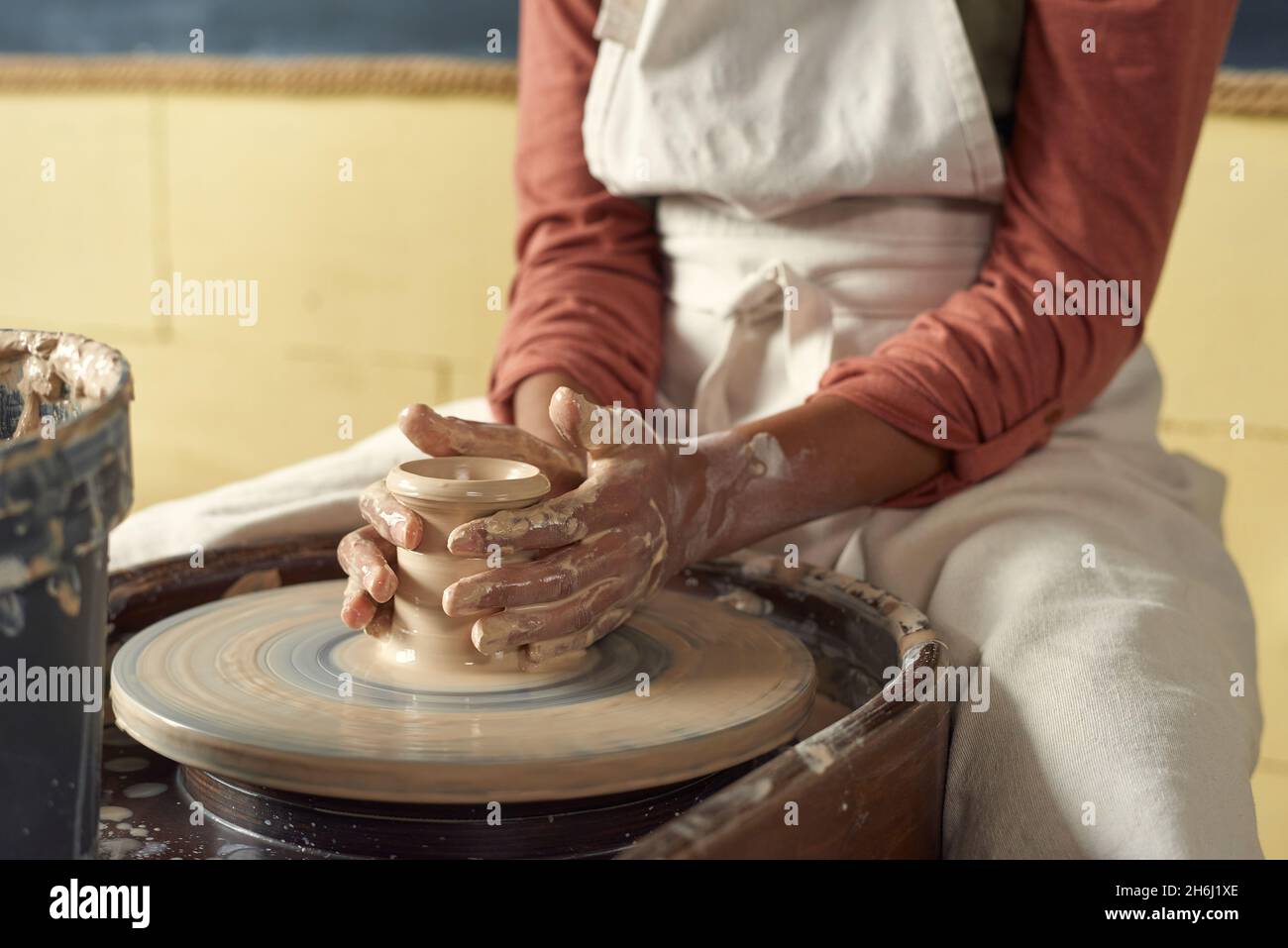 Schülerin in Schürze hält Tontopf auf rotierendem Töpferrad, während sie an einem Handwerkskurs oder Kurs teilnimmt Stockfoto
