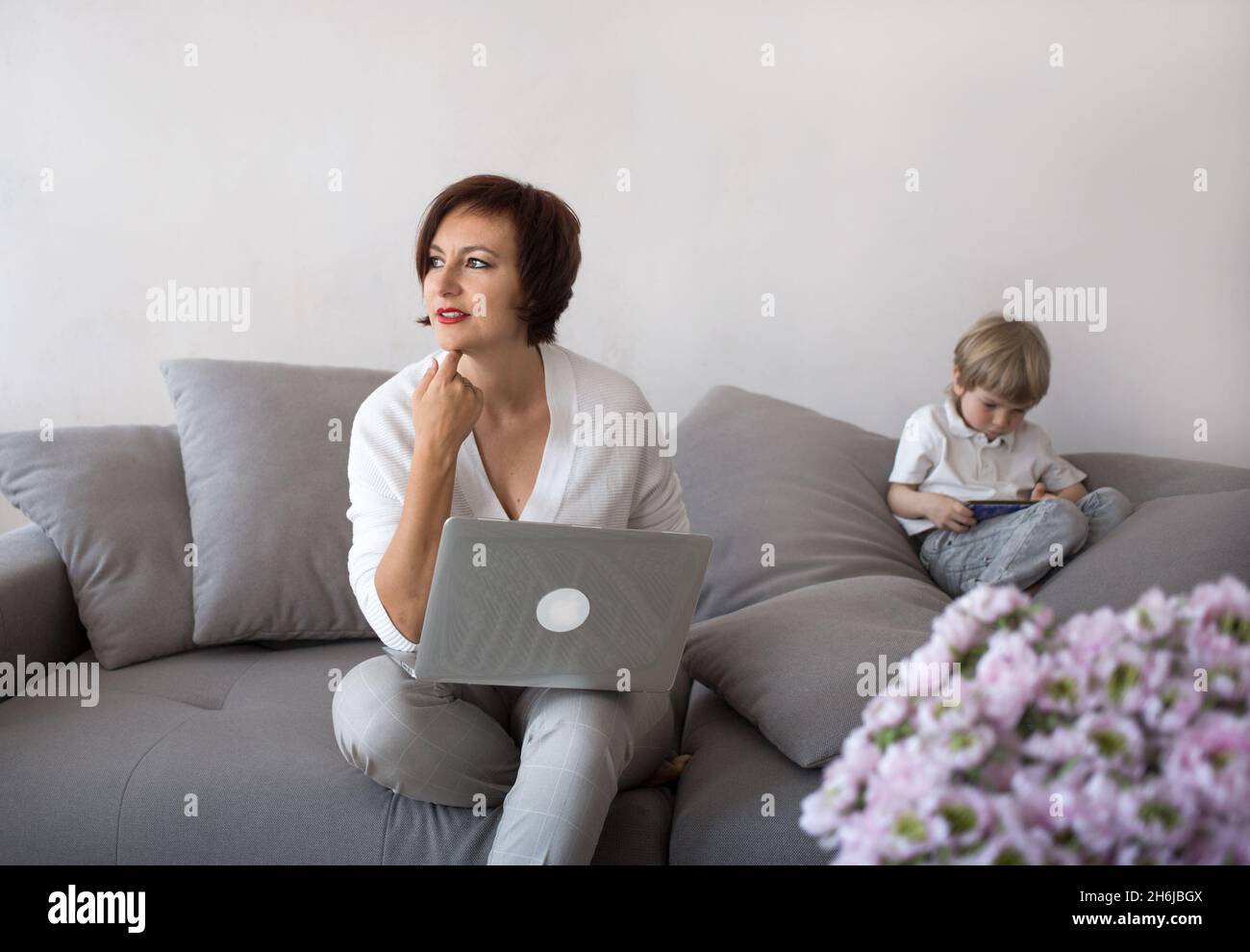 Mutter und Sohn mit digitalen Gadgets im Wohnzimmer. Eine Frau arbeitet zu Hause auf einem Laptop, neben einem Kind spielt am Telefon. Das Konzept der Moderne Stockfoto