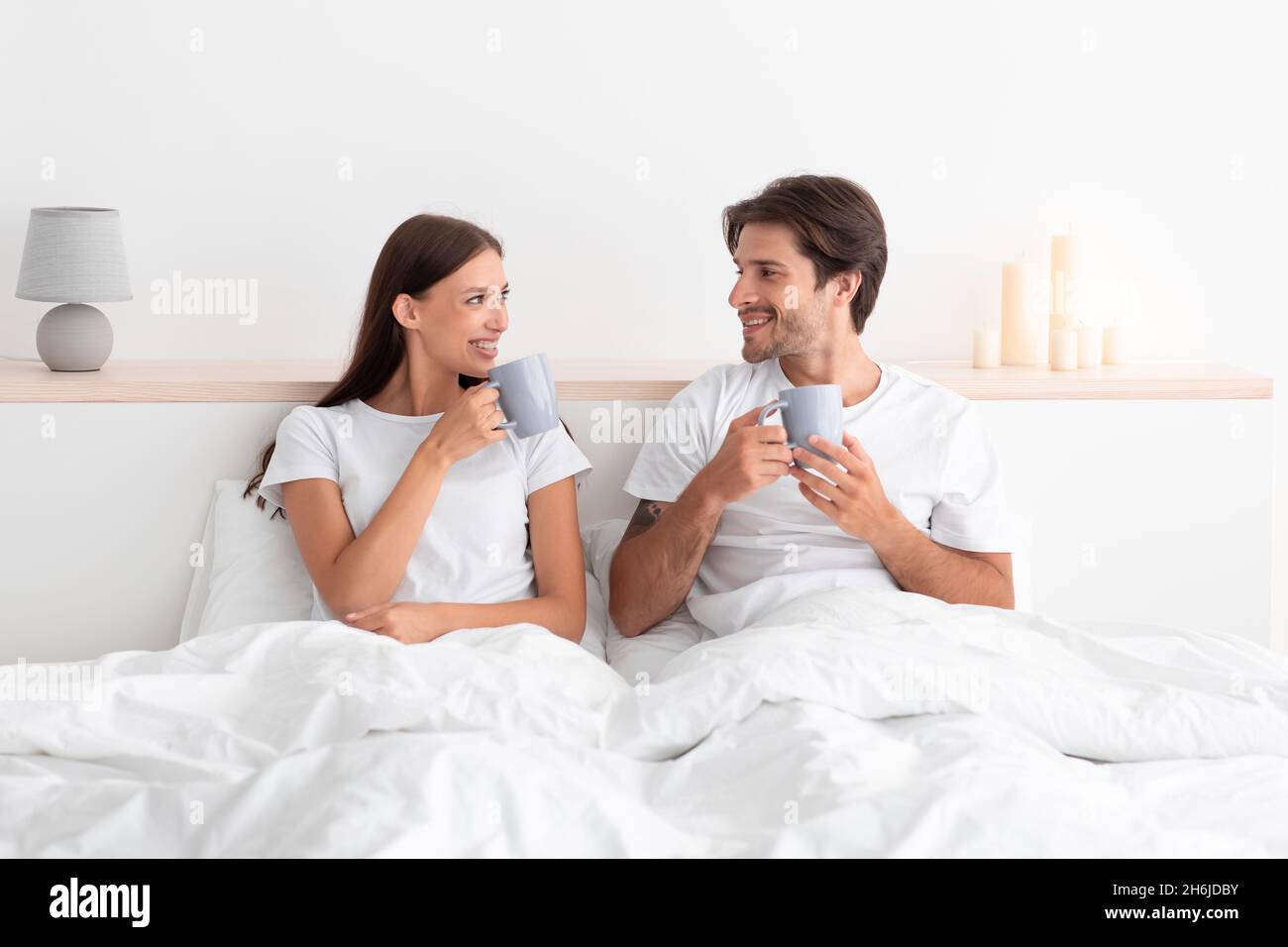 Lächelndes junges europäisches Paar wacht auf und genießt frischen Kaffee auf dem Bett im weißen Schlafzimmer Stockfoto