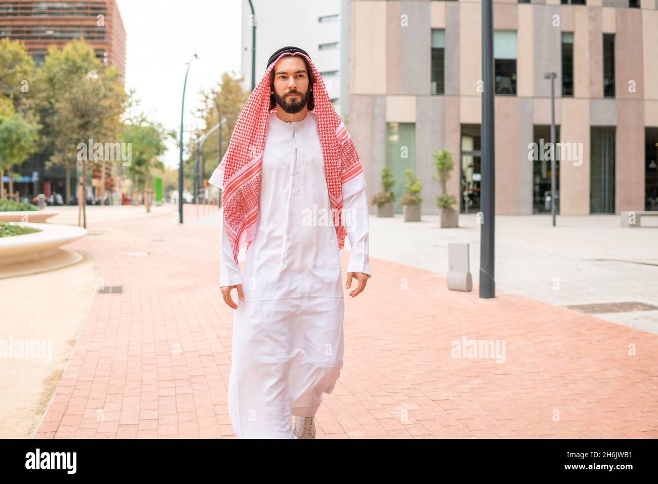 Gutaussehender erfolgreicher arabischer Mann sheikh in traditioneller Kleidung, der die Stadtstraße in Dubai zu Fuß führt, selbstbewusster arabischer Geschäftsmann in weißer saudischer Kandora im Freien Stockfoto