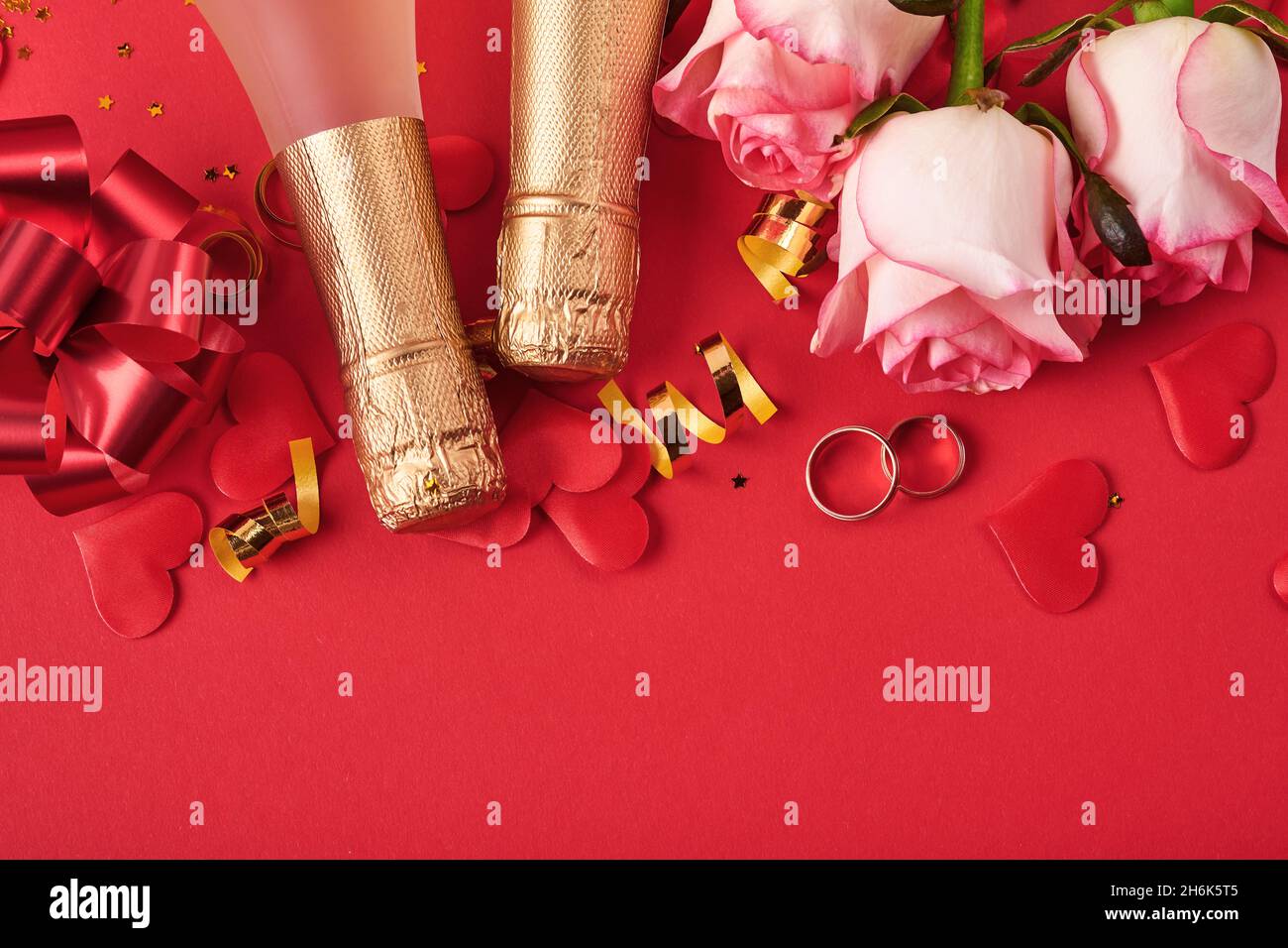 Rosa Rosen Blumen, Eheringe, Champagner, Geschenk, goldene Bänder und Konfetti Herzen auf rotem Hintergrund. Draufsicht mit Platz für Begrüßungen. Valentinstag Stockfoto