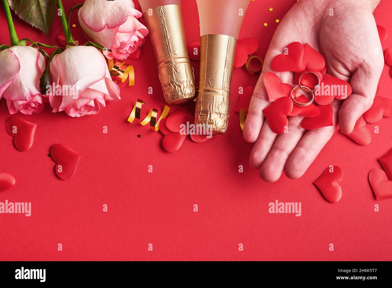 Rosa Rosen Blumen, Eheringe in männlichen Händen, Champagner, Geschenk, goldene Bänder und Konfetti Herzen auf rotem Hintergrund. Draufsicht. Valentinstag oder wir Stockfoto