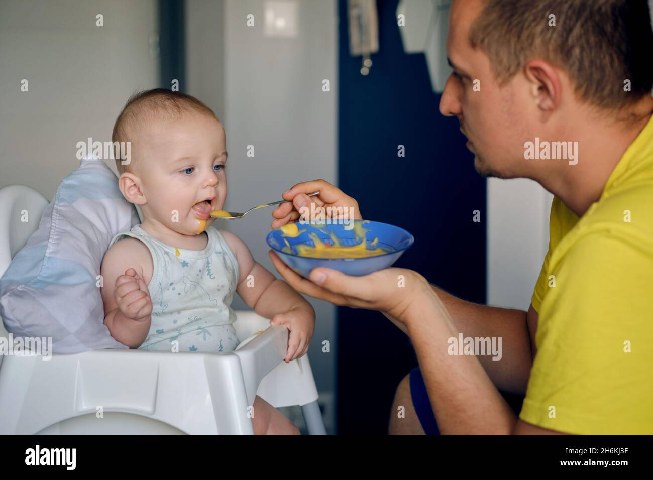 Ein junger Kaukasusmann füttert ein entzückendes blauäugig-blaues Baby mit gelber Babynahrung Stockfoto