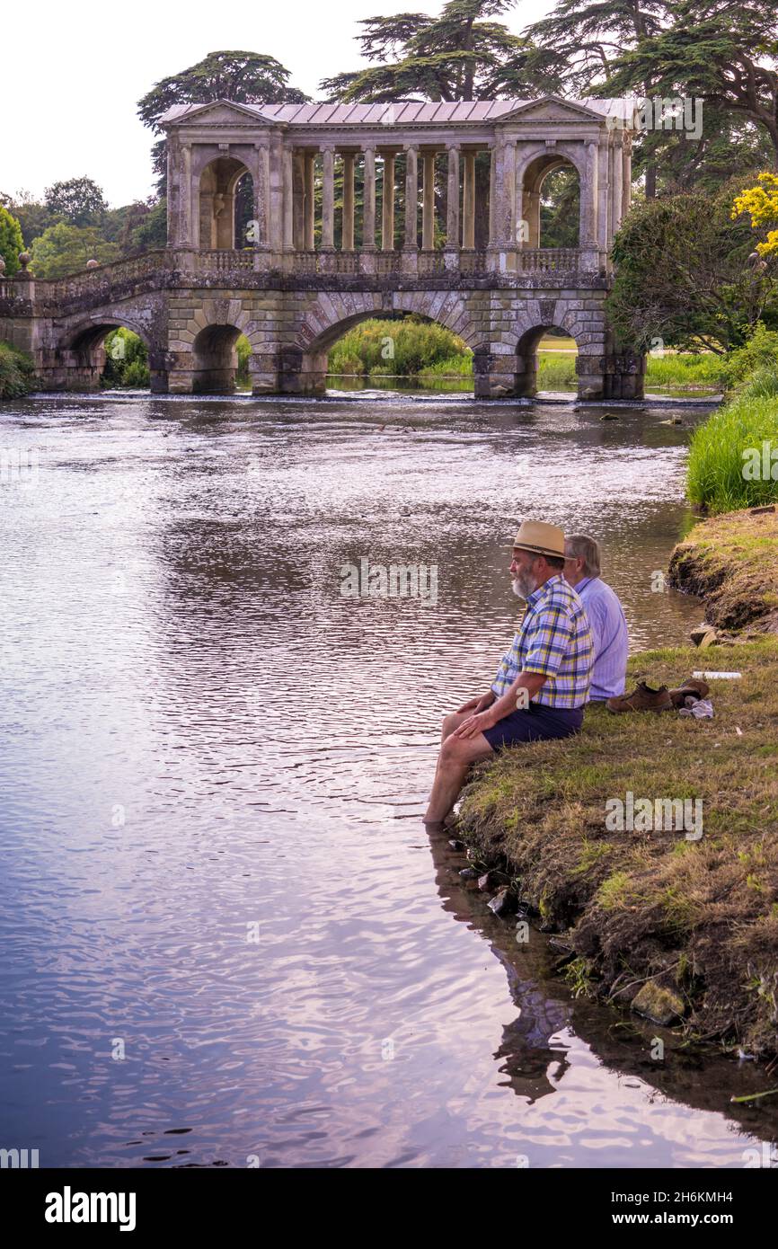 Zwei Menschen sitzen am Flussufer mit Füßen auf dem Wasser am Fluss Nadder mit den Palladian Bridge-Gärten und parken im Wilton House Wilton England Stockfoto