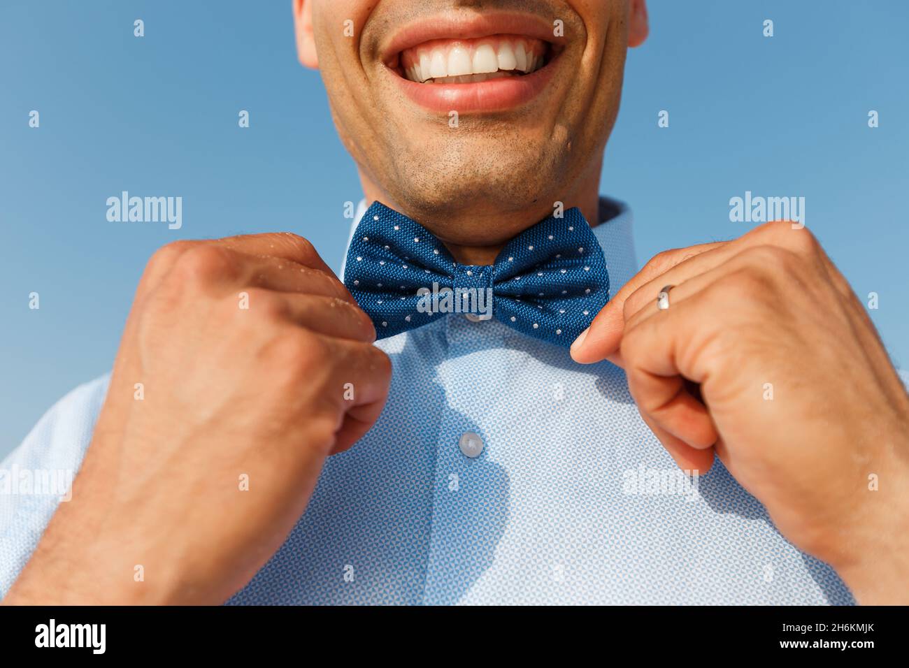 Junger lächelnder Mann in blauem Hemd berührt die Fliege auf dem Hintergrund des blauen Himmels. Accessoires und Lifestyle für Herren Stockfoto