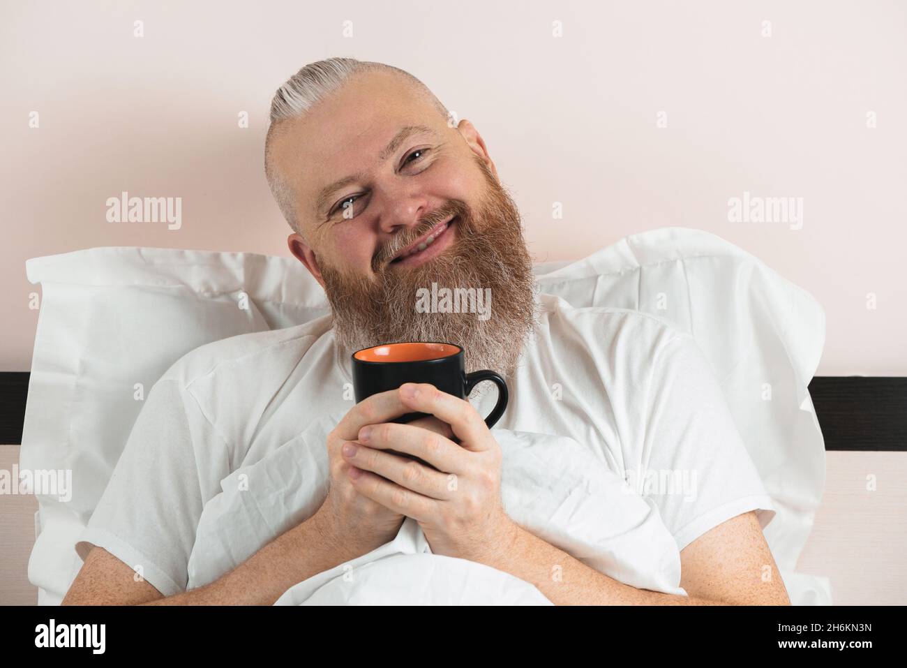 Glücklicher bärtiger Mann, der morgens Kaffee im Bett genießt. Lächelnder Hipster-Typ beginnt den Tag. Nahaufnahme. Stockfoto