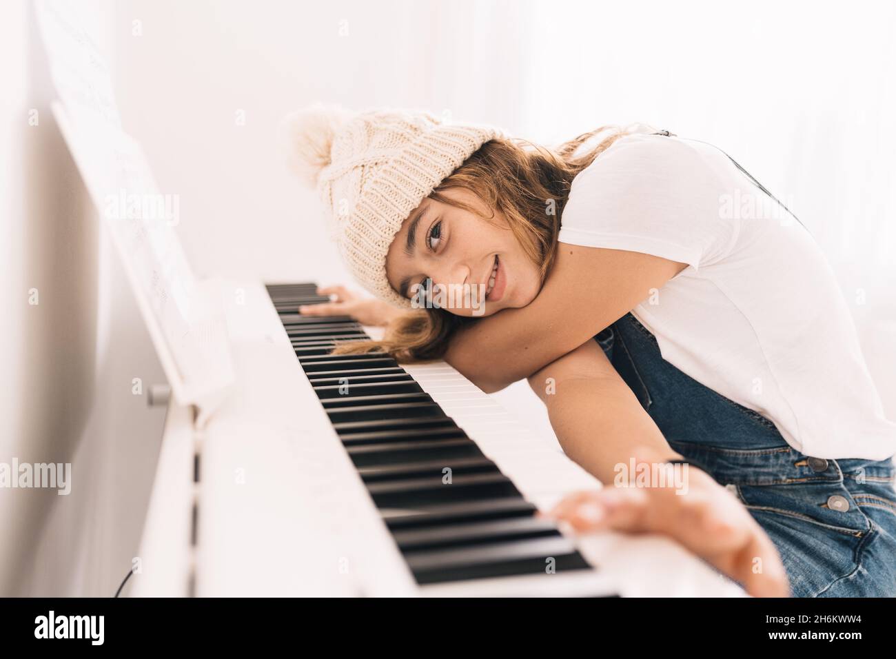 Teenager-Mädchen in Jeans und Wollmütze, die in ihrem Zimmer das weiße Klavier spielen, während sie die Musik auf pinken Kopfhörern hören. Weiches natürliches Nachhall Stockfoto