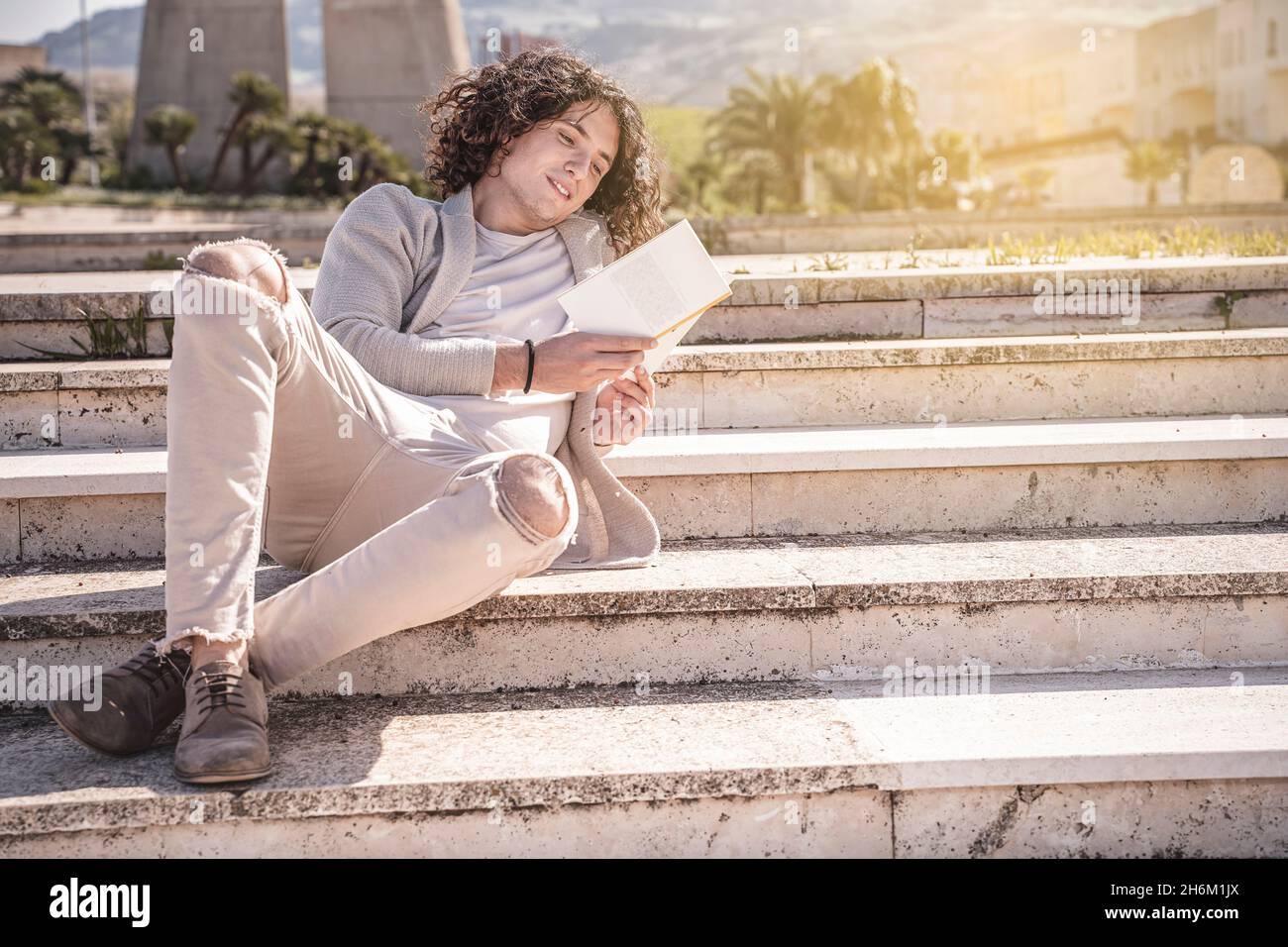 Ein junger Mann, der auf der Treppe sitzt, liest in einer Städtereise im Freien ein Buch Stockfoto