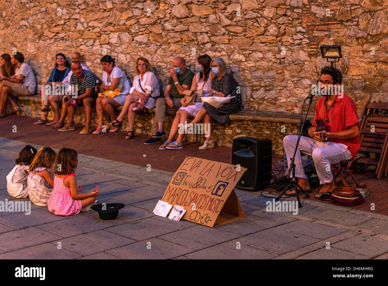 Ein Musiker mit Mikrofon spielt vor drei sitzenden Kindern in der spanischen Stadt Begur an der Costa Brava. Stockfoto