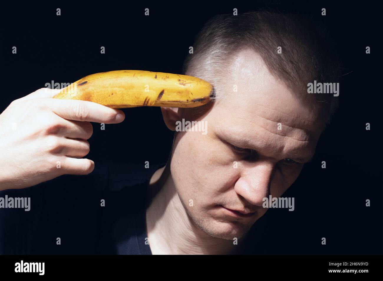 Der Mann hält Banane wie eine Pistole auf schwarzem Hintergrund nahe an seinem Kopf. Schießen Sie sich mit Banane. Verzweiflung, Depression und Selbstmordgedanken. Mental Stockfoto