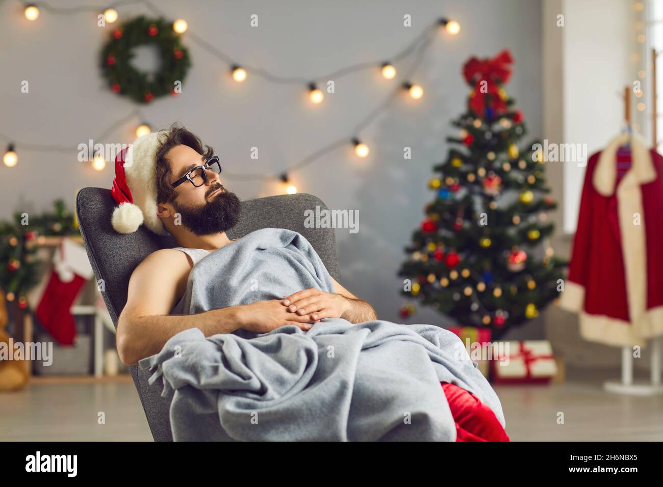 Bärtiger lustiger Mann in der Weihnachtsmütze, der unter der Decke im Sessel schläft Stockfoto