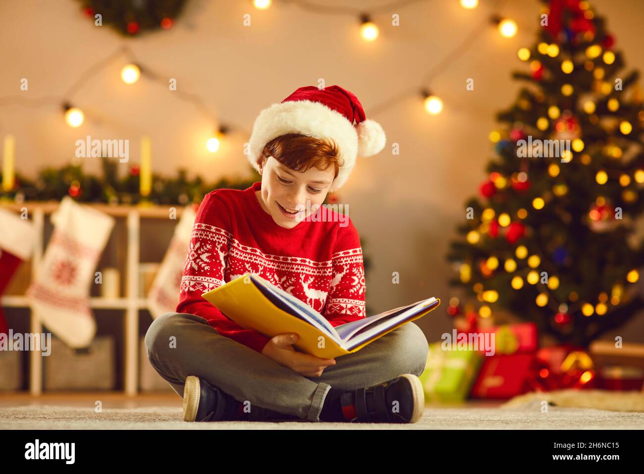 Positiver Junge mit Weihnachtsmütze und rotem traditionellem Pullover, der auf dem Boden sitzt und Buch liest Stockfoto