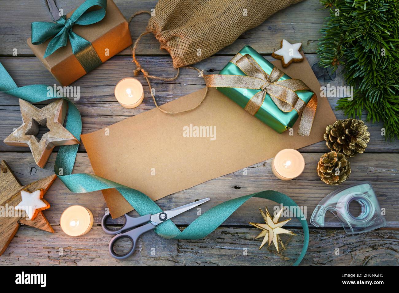 Geschenkverpackung mit braunem Papier und türkisfarbenem Band, Scheren, Dekorationssternen und brennenden Kerzen auf einem grauen rustikalen Holztisch, Kopierraum Stockfoto