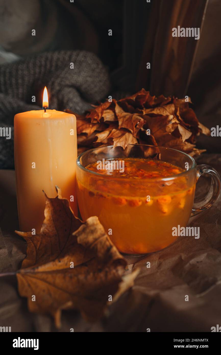 Brennende Kerze und eine große Tasse heißen orangefarbenen Sanddorntee in einem gemütlichen herbstlichen Stillleben mit trockenen Blättern. Stockfoto