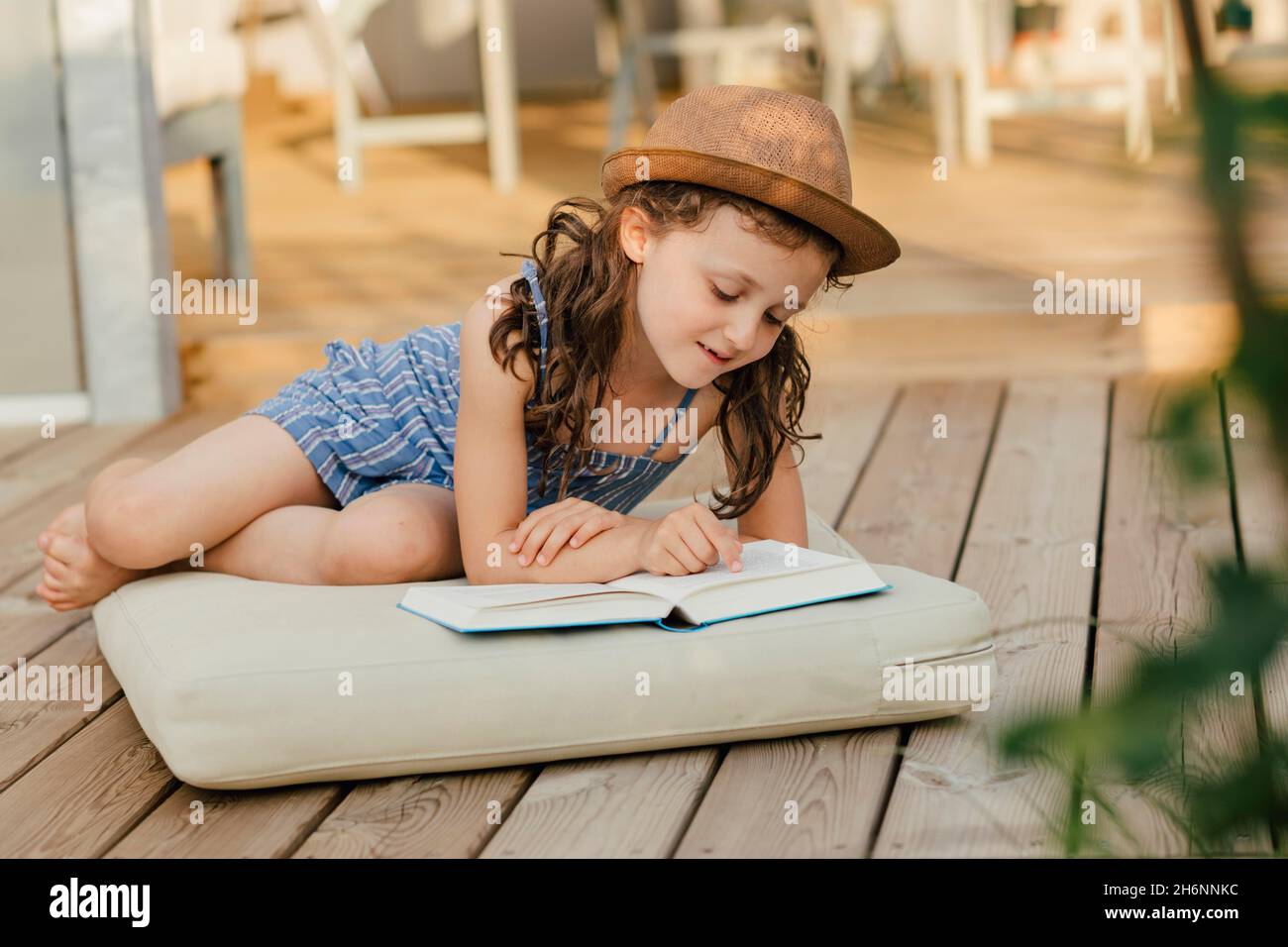 Mädchen, das auf dem Kissen auf der Holzterrasse sitzt und ein Buch liest Stockfoto